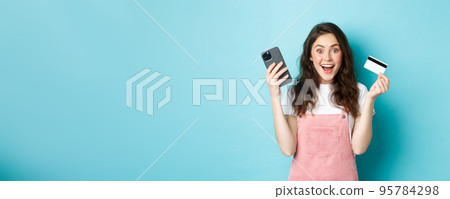 Cheerful girl looks excited at camera, hold smartphone and plastic credit card, standing over blue background Cheerful girl looks excited at camera, hold smartphone and plastic credit card, standing over blue background 95784298