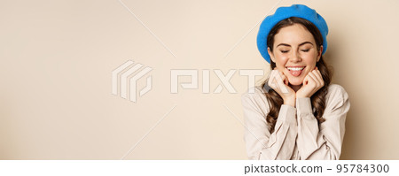 Close up portrait of feminine young woman in trendy french hat, posing cute and romantic, making silly coquettish face and tender smile, beige background Close up portrait of feminine young woman in trendy french hat, posing cute and romantic, making silly coquettish face and tender smile, beige background 95784300