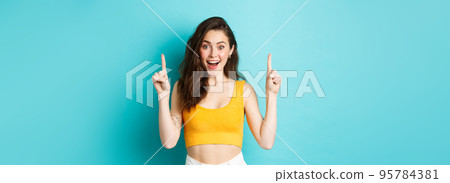 Summer lifestyle concept. Surprised happy woman showing advertisement on top, pointing fingers up and smiling excited, checking out special deal, standing over blue background 95784381
