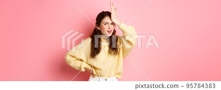 Annoyed and frustrated young woman face palm, holding hand on forehead and staring disappointed at camera, standing against pink background 95784383