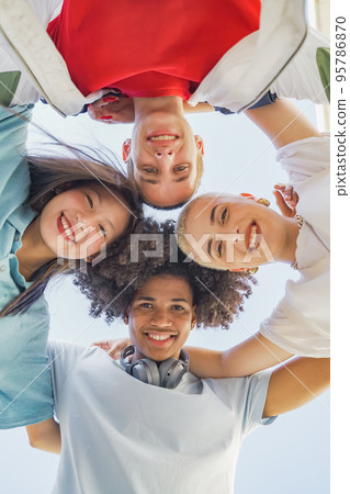 Low angle view of multiethnic young trendy friends hugging, smiling and making a circle looking at camera. 95786870