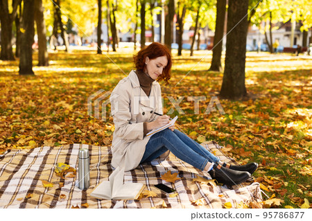 Concentrated smart young european red-haired lady student in raincoat sit with phone makes notes in diary 95786877