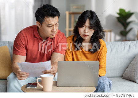 Upset asian couple checking bills, using laptop, home interior Upset asian couple checking bills, using laptop, home interior 95787465