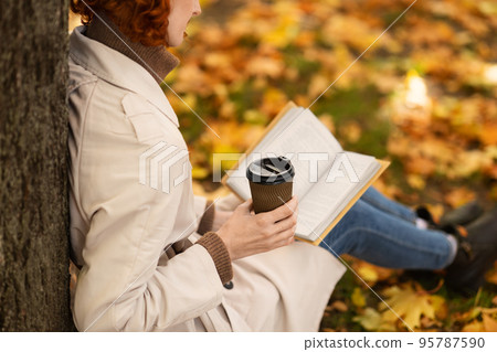 Young european red-haired woman with cup of drink takeaway in raincoat reads book near tree in park Young european red-haired woman with cup of drink takeaway in raincoat reads book near tree in park 95787590