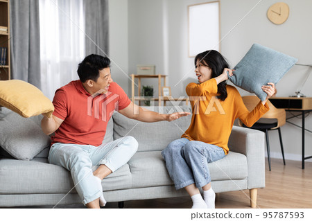 Asian spouses in homewear sitting on couch, having pillow fight Asian spouses in homewear sitting on couch, having pillow fight 95787593