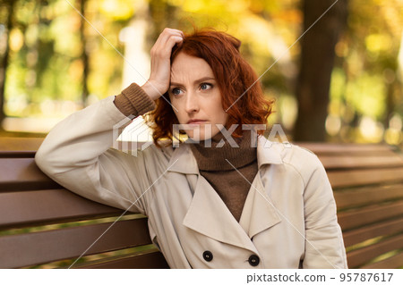 Despaired unhappy young european red-haired lady in raincoat suffers from headache, loneliness sits on bench Despaired unhappy young european red-haired lady in raincoat suffers from headache, loneliness sits on bench 95787617