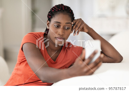 Pensive African American Female Looking At Smartphone Screen While Sitting At Home 95787817