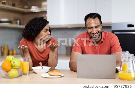 Sad dissatisfied young black woman resents and swears at smiling husband with pc at kitchen interior Sad dissatisfied young black woman resents and swears at smiling husband with pc at kitchen interior 95787926