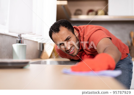 Sad dissatisfied young black male in red t-shirt wipes dirt from wooden table in kitchen interior, close up Sad dissatisfied young black male in red t-shirt wipes dirt from wooden table in kitchen interior, close up 95787929