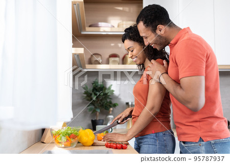 Smiling young black husband in red t-shirt hugs his wife, lady prepares food with organic vegetables Smiling young black husband in red t-shirt hugs his wife, lady prepares food with organic vegetables 95787937