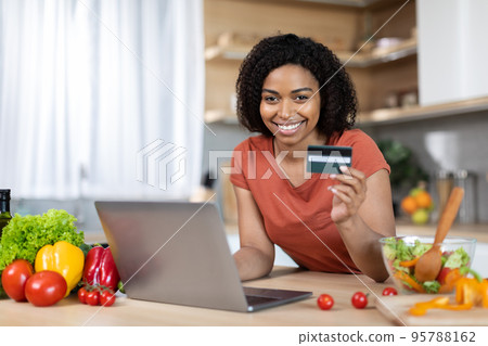 Smiling young black woman in red t-shirt with computer show credit card at table with vegetables Smiling young black woman in red t-shirt with computer show credit card at table with vegetables 95788162