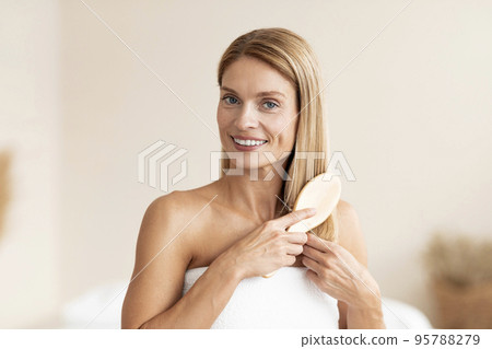 Morning beauty routine. Attractive mature woman brushing her blonde hair with comb, standing wrapped in bath towel Morning beauty routine. Attractive mature woman brushing her blonde hair with comb, standing wrapped in bath towel 95788279
