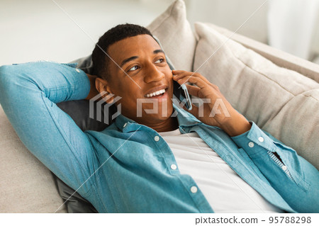 Smiling millennial african american man lying on sofa making phone call in living room interior, close up 95788298