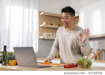 Asian man cooking dinner, having video conversation, using laptop Asian man cooking dinner, having video conversation, using laptop 95788356