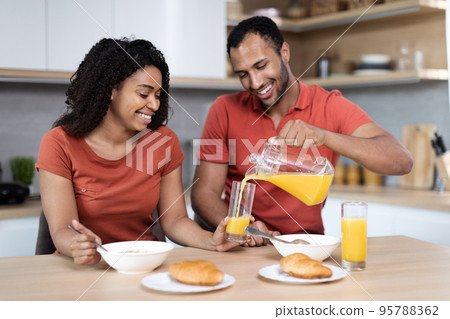Happy handsome young black guy in red t-shirt pours juice into glass to his wife, couple enjoys breakfast Happy handsome young black guy in red t-shirt pours juice into glass to his wife, couple enjoys breakfast 95788362