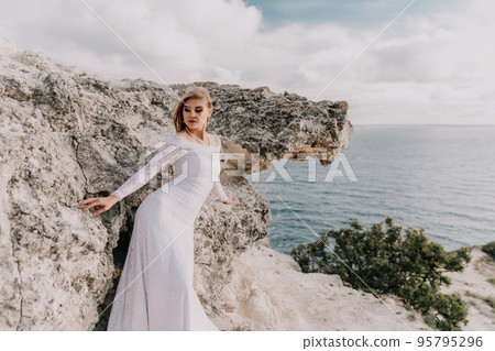 Romantic bride, blond girl in white wedding dress with open shoulders posing with sea and rocks in background. Stylish young woman standing on cliff's edge and looking into a ocean mountains at sunset Romantic bride, blond girl in white wedding dress with open shoulders posing with sea and rocks in background. Stylish young woman standing on cliff's edge and looking into a ocean mountains at sunset 95795296