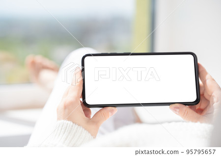 mockup cellphone. Woman lies in relaxed position and watches video content on cellphone or mobile smartphone with white blank screen. holding phone horizontally 95795657