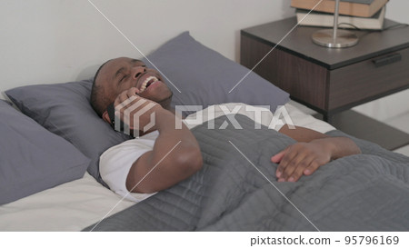 African Man Talking on Smartphone while Sleeping in Bed, Close up African Man Talking on Smartphone while Sleeping in Bed, Close up 95796169
