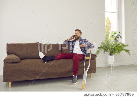 Happy man with broken leg talking to someone on phone while sitting on couch at home Happy man with broken leg talking to someone on phone while sitting on couch at home 95797999