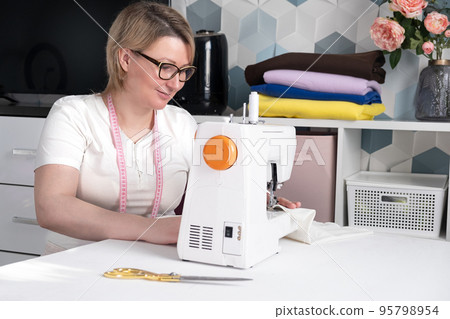 Real middle aged woman tailor sewing clothes using a sewing machine at home. Seamstress working. Hand making clothes in home interior. Female sits enjoying process of sewing clothes. Real middle aged woman tailor sewing clothes using a sewing machine at home. Seamstress working. Hand making clothes in home interior. Female sits enjoying process of sewing clothes. 95798954