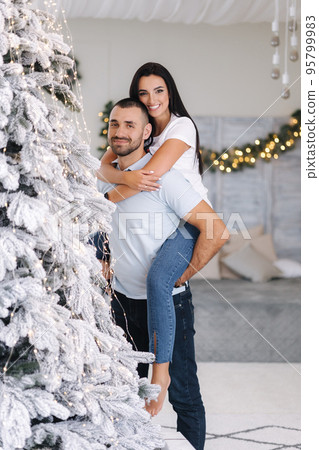 Beautiful woman climbed behind her husband's back and hugged him. Cute couple spend time at home during Christmas. Fit tree. Fairytale 95799983