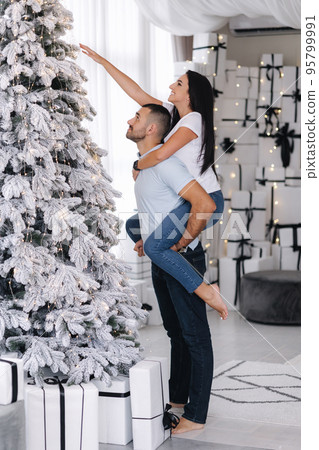Beautiful woman climbed behind her husband's back to decorate Christmas tree. Cute couple spend time at home during holidays 95799991