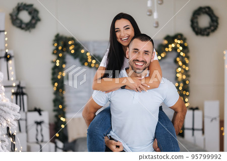 Beautiful woman climbed behind her husband's back and hugged him. Cute couple spend time at home during Christmas. Fit tree Beautiful woman climbed behind her husband's back and hugged him. Cute couple spend time at home during Christmas. Fit tree 95799992