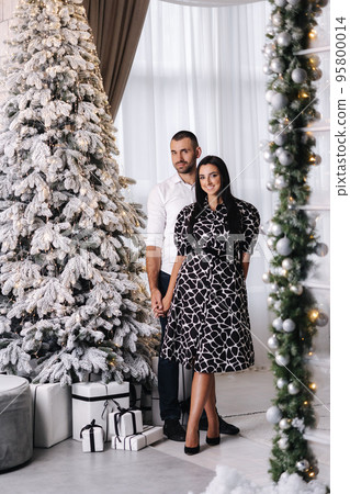 Attractive female in beautiful black and white dress stand with her husband in front of fit tree. Christmas mood at home Attractive female in beautiful black and white dress stand with her husband in front of fit tree. Christmas mood at home 95800014