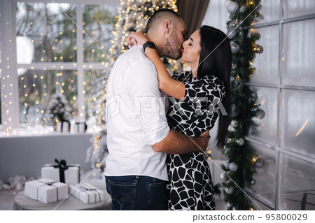 Handsome man with his beautiful woman hug each other at home at Christmas eve. Fit tree and snow outsif. Christmas mood Handsome man with his beautiful woman hug each other at home at Christmas eve. Fit tree and snow outsif. Christmas mood 95800029