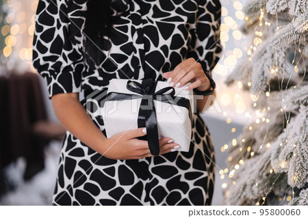 Middle selection of brunette woman in dress open present on Chrisrmas time. Elegant woman stand by big Christmas tree in front of window Middle selection of brunette woman in dress open present on Chrisrmas time. Elegant woman stand by big Christmas tree in front of window 95800060