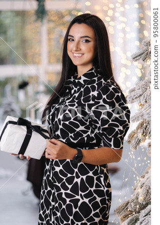 Portrait of brunette woman in dress open present on Chrisrmas time. Elegant woman stand by big Christmas tree in front of window Portrait of brunette woman in dress open present on Chrisrmas time. Elegant woman stand by big Christmas tree in front of window 95800061