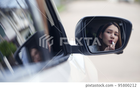 Young woman applying lipstick looking at reflection in car mirror. 95800212