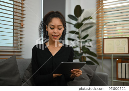 Beautiful young African woman relaxing on sofa in cozy living room and using digital tablet 95800521