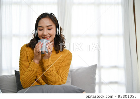 Relaxed Asian woman listening to music while sipping hot coffee or tea in her living room. Relaxed Asian woman listening to music while sipping hot coffee or tea in her living room. 95801053