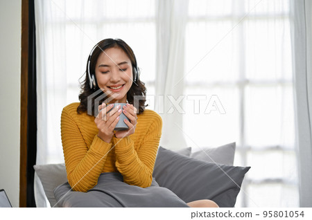 Pretty Asian woman listening to music while sipping her tea or coffee in the living room. 95801054