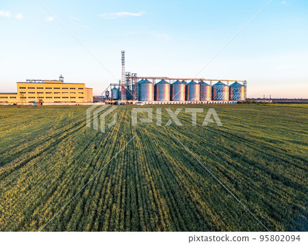 Grain elevator. Metal grain elevator in agricultural zone. Agriculture storage for harvest. Grain silos on green nature background. Exterior of agricultural factory. Sunset warm light. Nobody. 95802094