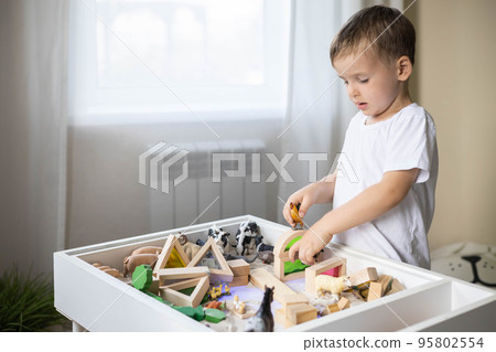 Smiling boy kid playing sensory box kinetic sand table with Montessori wooden materials Smiling boy kid playing sensory box kinetic sand table with Montessori wooden materials 95802554