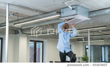 Caucasian bearded man repairing the air conditioner in the office. Caucasian bearded man repairing the air conditioner in the office. 95803607