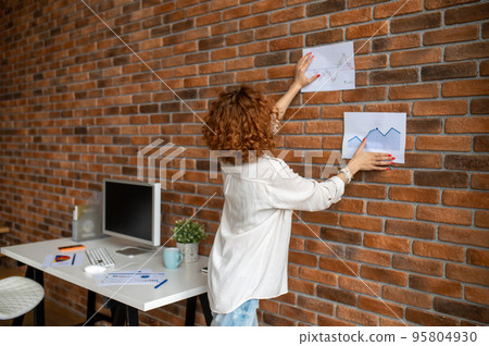 Ginger woman putting diagrams on the wall in the office Ginger woman putting diagrams on the wall in the office 95804930