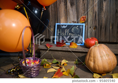 Halloween trick and treat. Still life. A whole pumpkin, candies and sweets in violet basket, orange and black air balloons and blackboard on the wooden threshold with fallen autumn dry colorful leaves Halloween trick and treat. Still life. A whole pumpkin, candies and sweets in violet basket, orange and black air balloons and blackboard on the wooden threshold with fallen autumn dry colorful leaves 95805540