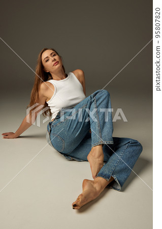 Portrait of beautiful young woman posing in casual clothes, jeans and white top over dark grey studio background. Stylish 95807820