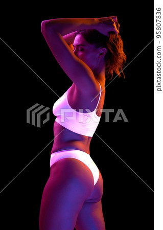 Portrait of beautiful woman with slim body posing in white underwear isolated over black background in neon light 95807836