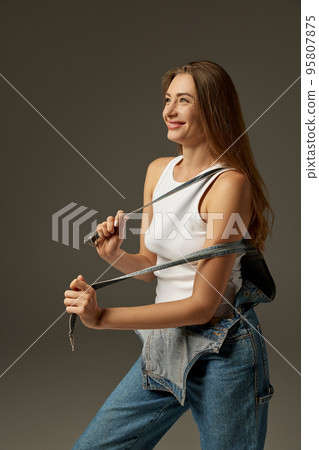 Portrait of beautiful cheerful woman posing in casual clothes, jeans and white top over dark grey studio background 95807875