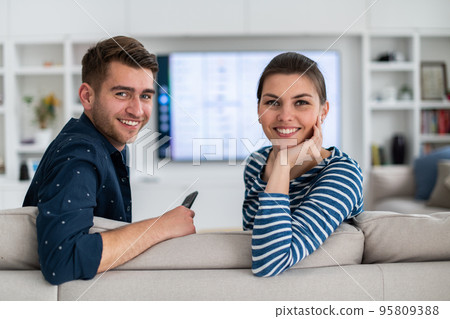 Couple Watches TV together while Sitting on a Couch in the Living Room. Girlfriend and Boyfriend embrace, cuddle, talk, smile and watch Television Streaming Services. Home with Cozy Stylish Interior. Couple Watches TV together while Sitting on a Couch in the Living Room. Girlfriend and Boyfriend embrace, cuddle, talk, smile and watch Television Streaming Services. Home with Cozy Stylish Interior. 95809388