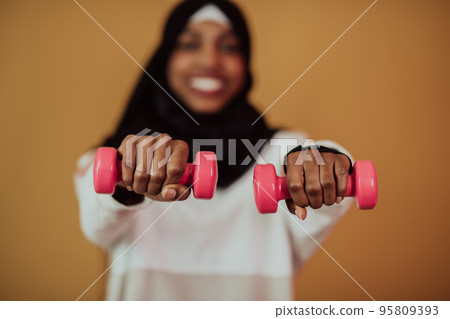 Afro muslim woman promotes a healthy life, holding dumbbells in her hands 95809393