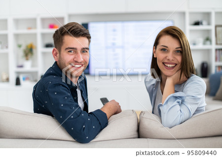 Couple Watches TV together while Sitting on a Couch in the Living Room. Girlfriend and Boyfriend embrace, cuddle, talk, smile and watch Television Streaming Services. Home with Cozy Stylish Interior. 95809440