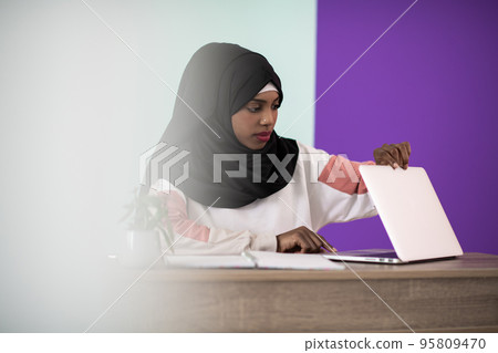 afro girl wearing a hijab is disappointed and sad sitting in her home office and using a laptop 95809470