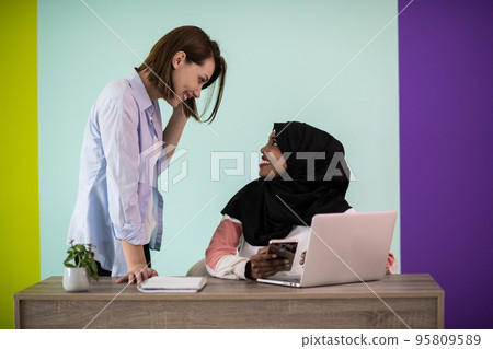 Afro woman with a hijab and a European woman using a smartphone and laptop in their home office 95809589