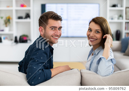 Couple Watches TV together while Sitting on a Couch in the Living Room. Girlfriend and Boyfriend embrace, cuddle, talk, smile and watch Television Streaming Services. Home with Cozy Stylish Interior. Couple Watches TV together while Sitting on a Couch in the Living Room. Girlfriend and Boyfriend embrace, cuddle, talk, smile and watch Television Streaming Services. Home with Cozy Stylish Interior. 95809762