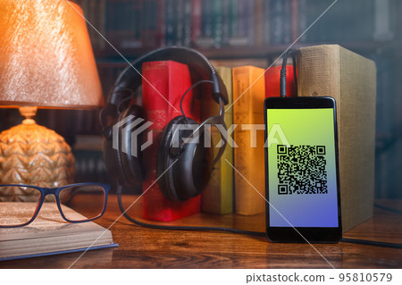 On a wooden table next to the lamp are headphones, a phone, a stack of books and glasses. The concept of modern technology and audiobooks. QR-code On a wooden table next to the lamp are headphones, a phone, a stack of books and glasses. The concept of modern technology and audiobooks. QR-code 95810579